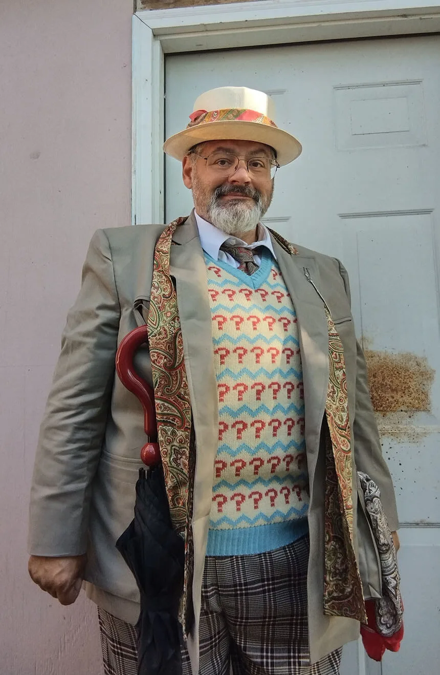 A man dressed as the Seventh Doctor from Doctor Who, standing in front of a door. He is wearing a stone colored blazer, a vest with red question marks, plaid trousers, a patterned scarf, and a hat. He holds a red-handled umbrella and is smiling at the camera.