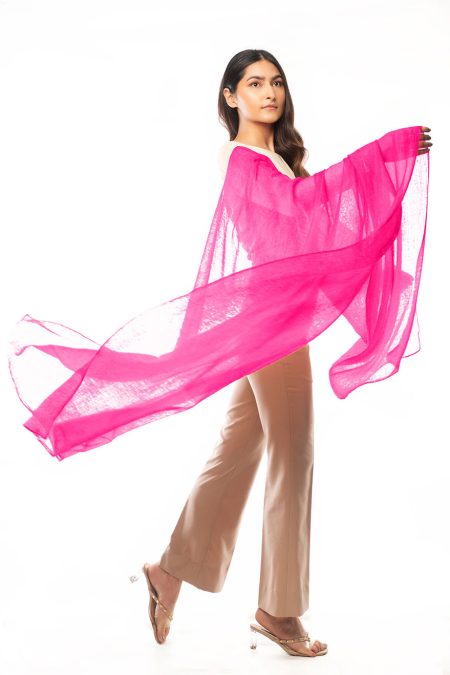 A woman drapes a bright pink cashmere scarf over her arm, highlighting its featherweight silhouette. She is wearing cream-colored top and beige high-waisted trousers.