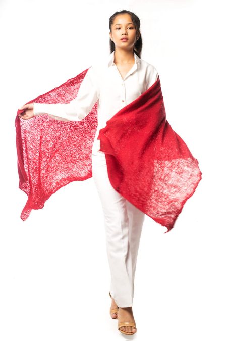 Model walking confidently, holding one end of a red cashmere scarf in one hand while spreading the other end to showcase the texture, width, and elegance of the hand-knitted 100% cashmere. The model is dressed in a white pant and shirt set, emphasizing the scarf's luxurious quality.