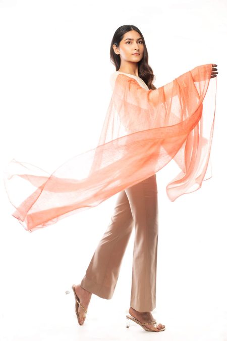 A woman drapes a salmon pink cashmere scarf over her arm, highlighting its airy silhouette. She is wearing cream-colored top and beige high-waisted trousers.