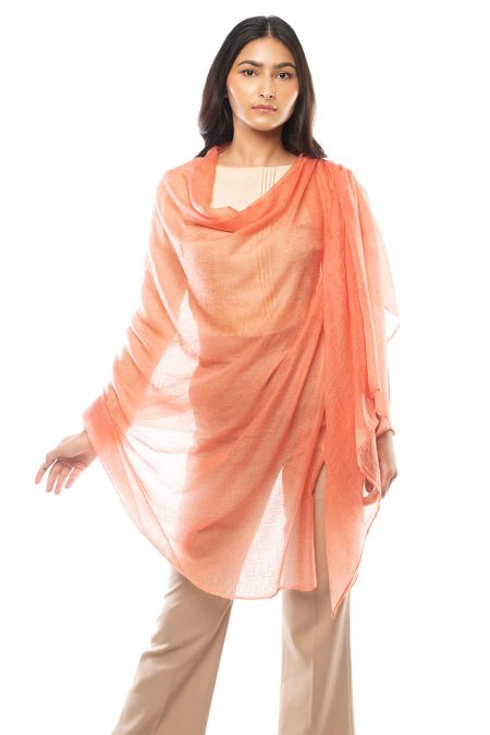 A woman wearing a salmon pink cashmere scarf elegantly draped around her body, paired with a beige top and tan high-waisted pants.