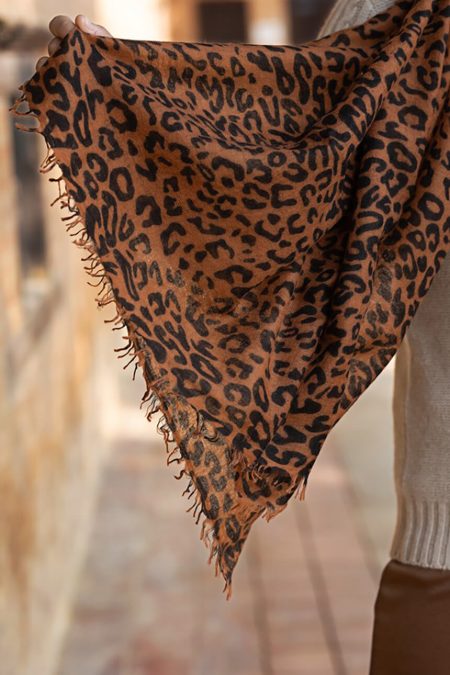 Hand holding a lightweight leopard print cashmere scarf with frayed edges, woven in twill weave, displayed against a warm-toned outdoor background.