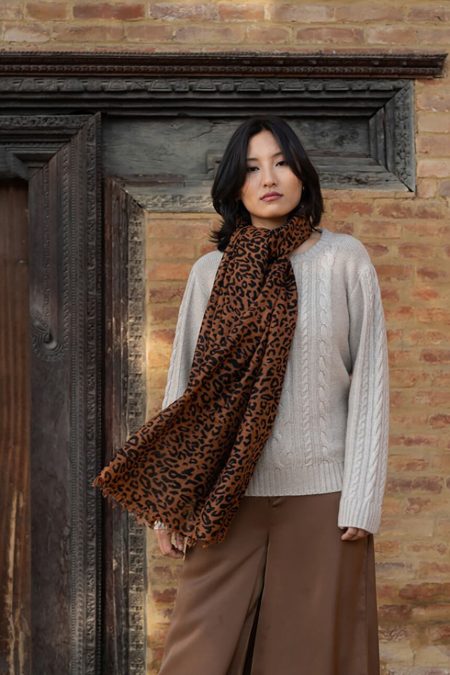 Woman wearing a leopard print cashmere scarf in a twill weave, styled over a cable-knit sweater standing against a rustic brick wall, while wind blows the cashmere scarf's edges.