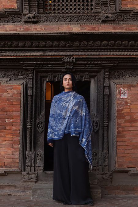 Woman wrapped in a blue and white cashmere scarf with antique landscape print, standing in front of a carved wooden doorway in Nepal.