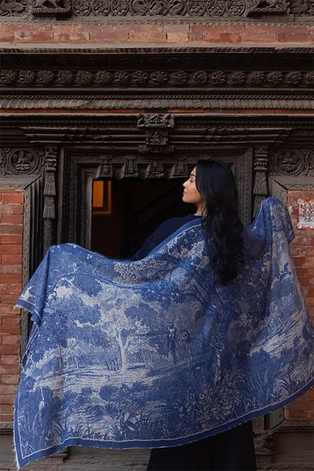 Woman draped in a blue and white cashmere scarf with antique landscape print, standing in front of a carved wooden doorway in Nepal.