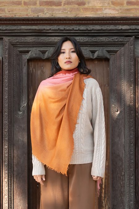 Woman wearing an orange rust ombre tie-dye cashmere scarf draped over a beige cable-knit sweater and brown trousers, standing against a carved wooden door with a brick wall backdrop — luxury lightweight twill weave scarf styled for autumn fashion.