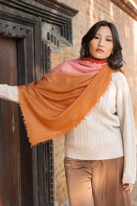 A stylish woman stands in front of an ornate wooden doorway, wearing a cream cable knit sweater, brown satin pants, and a pink to orange ombré cashmere scarf draped elegantly over her shoulder. Outdoor portrait highlighting the scarf and architectural details.