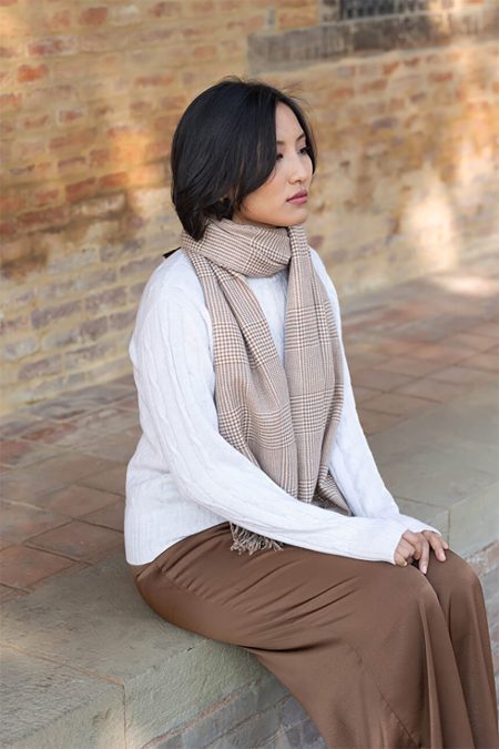 A woman sitting outside, wearing a tan and white glen plaid 100% cashmere scarf draped over a white sweater and brown pants.