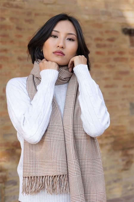 A woman standing and adjusting a tan and white glen plaid 100% cashmere scarf styled around her neck.