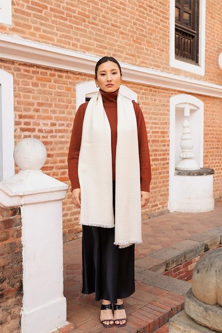 Woman models an off-white pearl weave cashmere scarf draped evenly over a rust-colored turtleneck sweater and black satin skirt, standing in front of a traditional brick building.