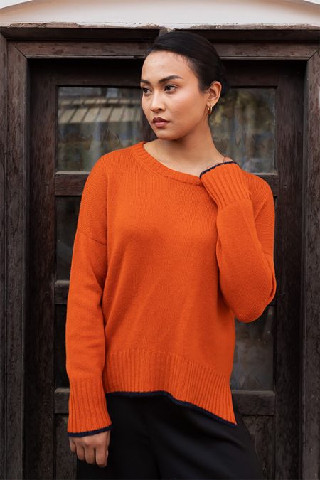 Three-quarter view of a woman wearing Baron Boutique's Ember Glow orange cashmere dropped shoulder boxy sweater, highlighting its contemporary boxy cut, round neck, dropped shoulder seams, and navy tipping detail.