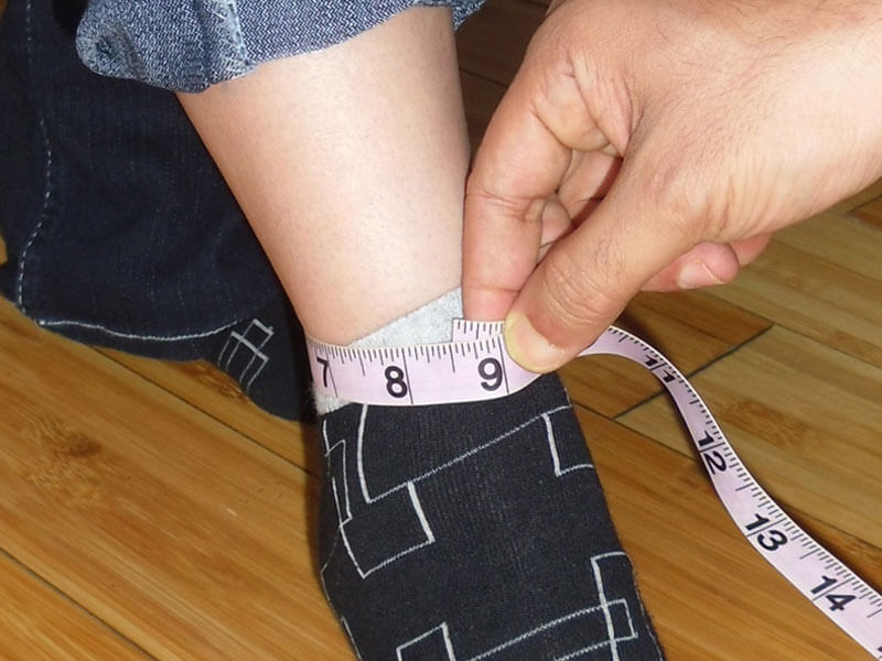 A close-up image demonstrating how to properly measure the ankle circumference for custom-tailored women's garments. A pink measuring tape is wrapped snugly around the ankle over a sock, showing a measurement of approximately 9 inches.