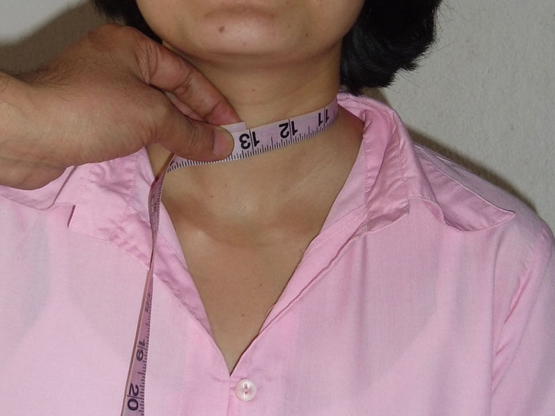 A close-up view of a neck measurement being taken for a custom-tailored women's collared shirt. The measuring tape is wrapped comfortably around the base of the neck to ensure the collar fits perfectly without being too tight.