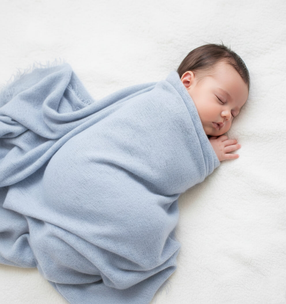 A peaceful newborn baby sleeping soundly, swaddled in an incredibly soft, hand felted powder blue cashmere blanket. The perfect heirloom gift for a baby shower.