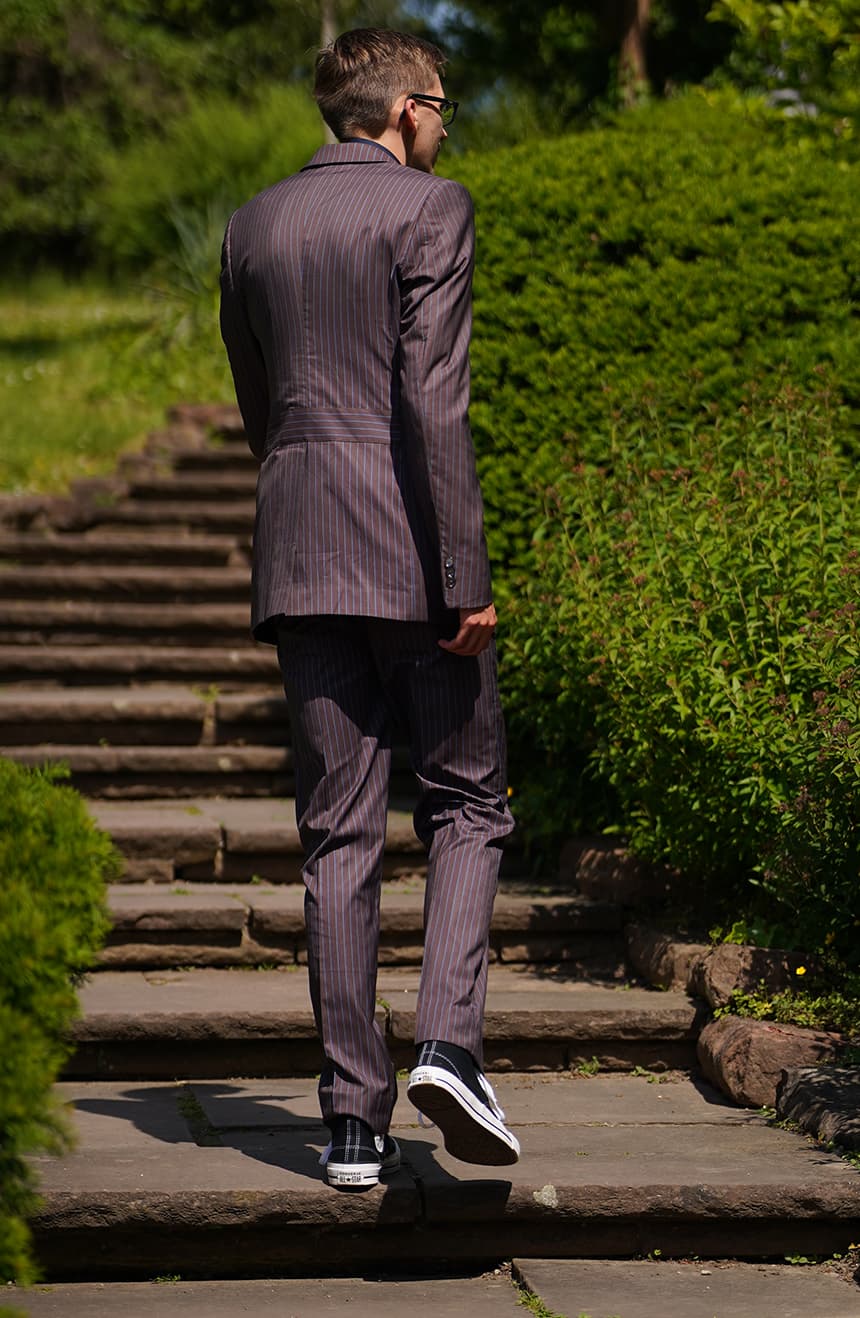 Man in a tenth Doctor brown pinstripe two-piece suit seen from behind walking up stone garden steps with black Converse sneakers.