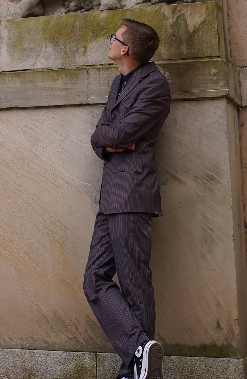 Side view of the Tennant brown and blue pinstripe suit on a man, highlighting the slim tailored silhouette, pocket details, subtle blue pinstripe accents, and elegant side profile against a sandstone wall.