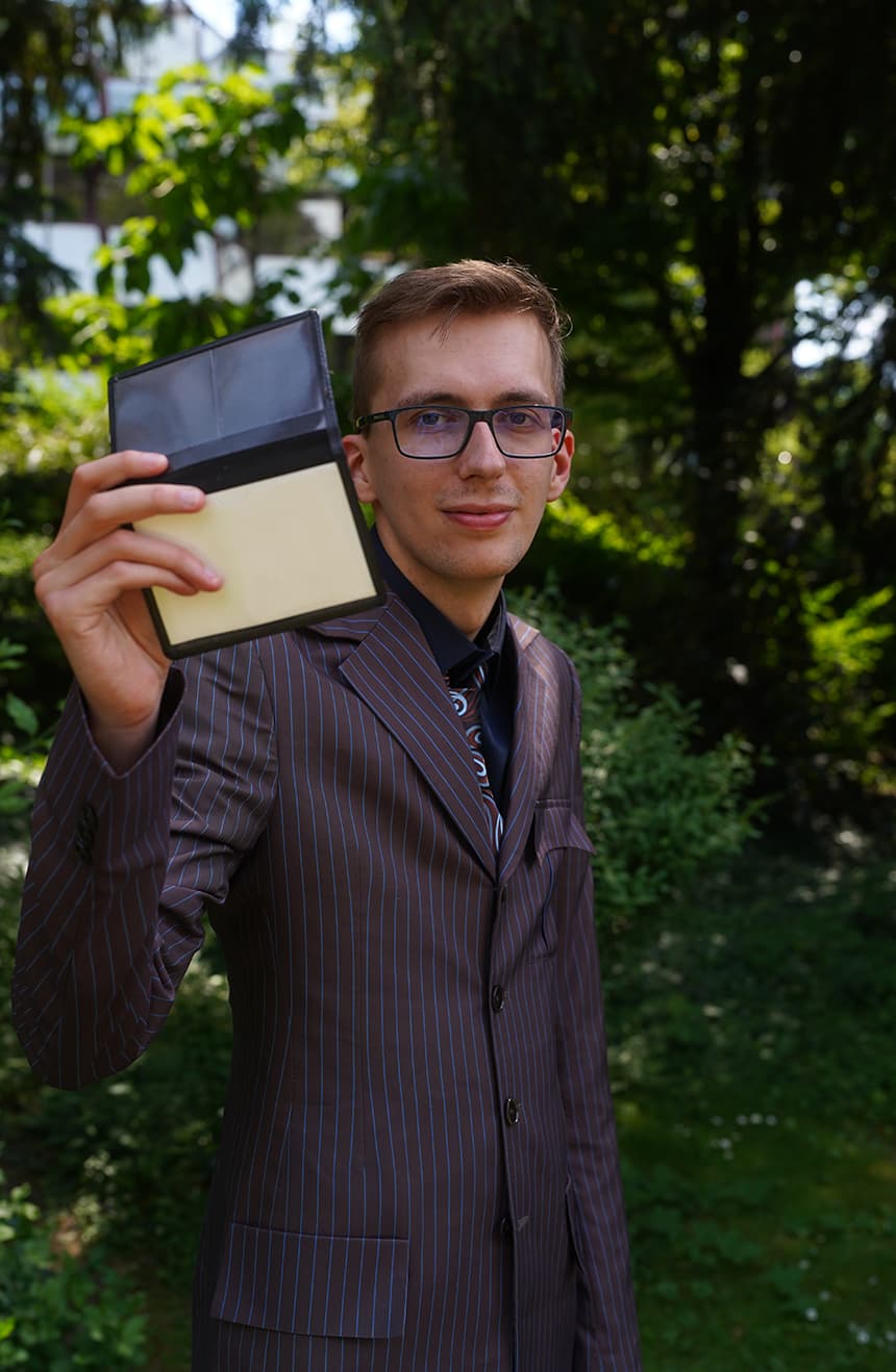 Man in a tenth brown with blue-pinstripe suit smiling and holding up a leather wallet in a green garden setting.