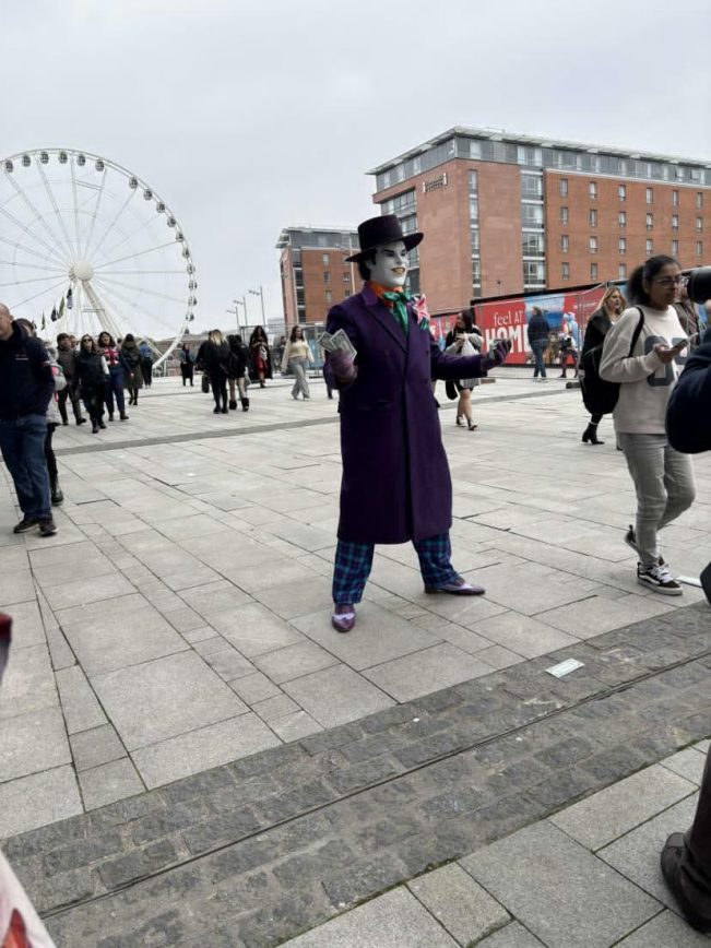 Full-length view of a customer modeling the custom 1989 purple coat outdoors, styled with a top hat, plaid trousers, and white clown face paint while holding prop money, set against a city plaza crowd with a large Ferris wheel in the background.