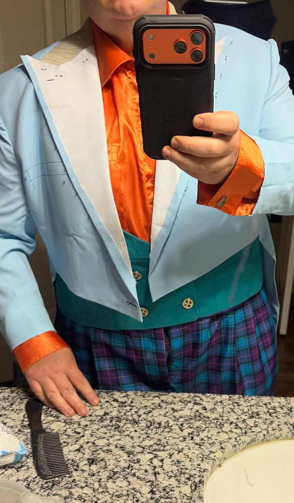 Mirror selfie of a customer modeling the light blue cotton mock-up test tailcoat, verifying fitment parameters and proportions over an orange shirt and plaid trousers before cutting the final luxury fabric.