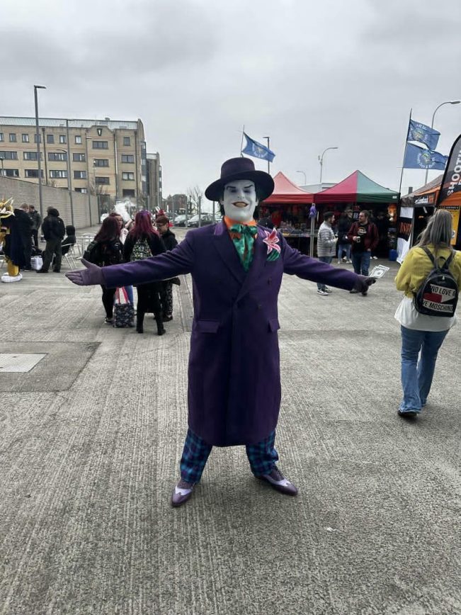 Full-length shot of a customer modeling the bespoke 1989 purple coat with open arms at an outdoor convention, styled with a black top hat, matching gloves, a floral lapel pin, and theatrical white face paint.