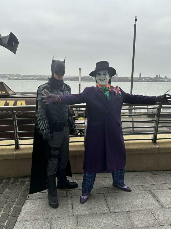 Wide full-length view of a customer modeling the bespoke 1989 purple coat replica with a top hat and white clown makeup, posing alongside a cosplayer in an armored bat vigilante suit by a waterfront boardwalk railing.
