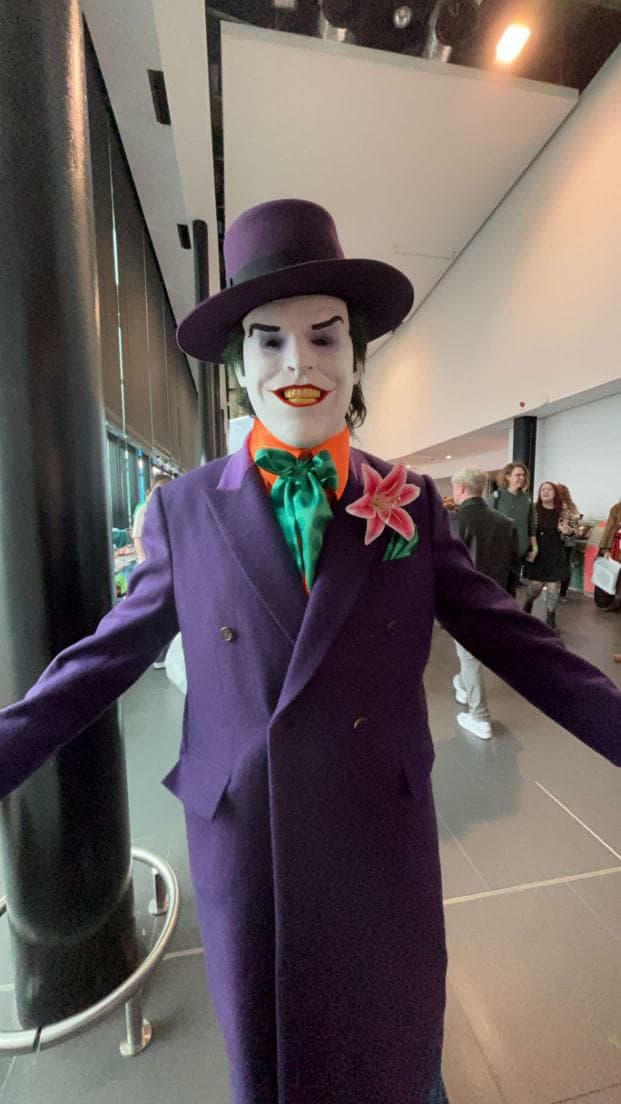 Close-up view of a happy customer modeling the custom 1989 purple coat replica with an orange silk shirt, sea green aviator neck scarf, matching purple top hat, and theatrical clown face paint inside a convention center hall.