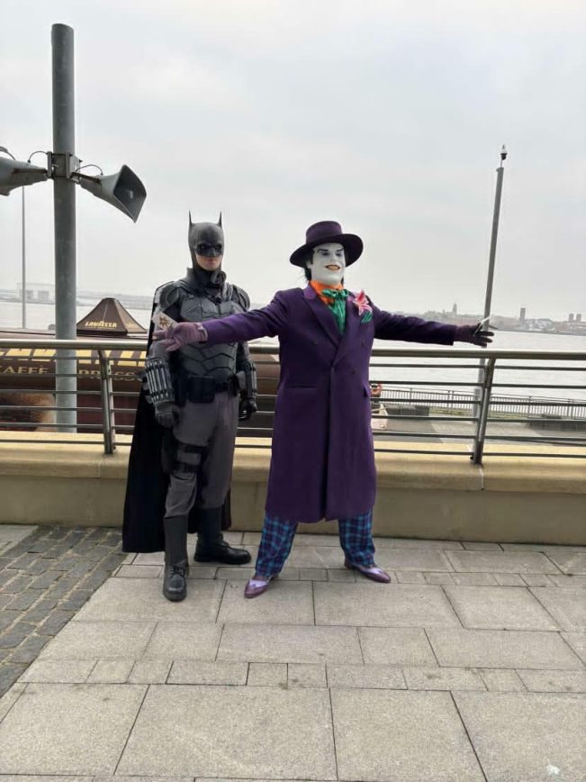 Full-length view of a customer wearing the custom-tailored 1989 purple coat posing with wide open arms alongside another enthusiast in a dark armored bat vigilante suit next to a waterfront railing.