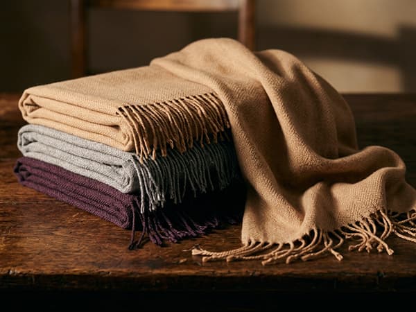 Handwoven cashmere scarves and a shawl in camel, grey and plum, folded to show the weave and fringed edge.