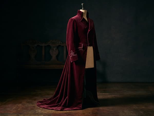 A burgundy velvet frock coat with a high collar and long tails on a dress form, lit theatrically.