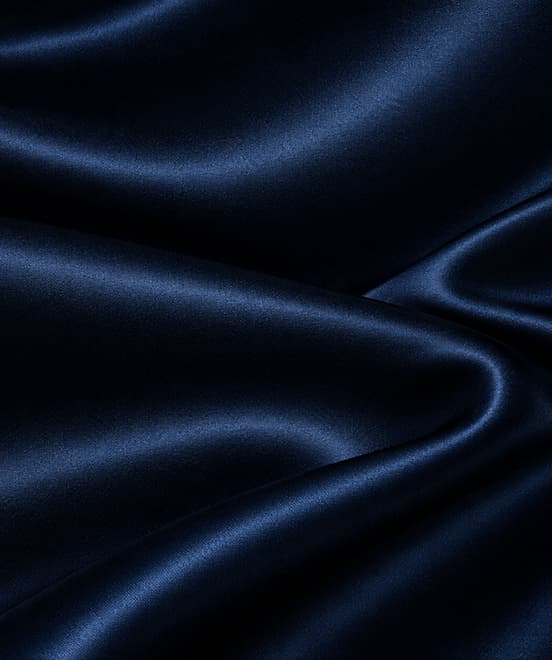 Close-up of heavy navy 22 momme mulberry silk charmeuse falling in deep folds, showing a soft sheen on the crest of each fold and a fully opaque weave.